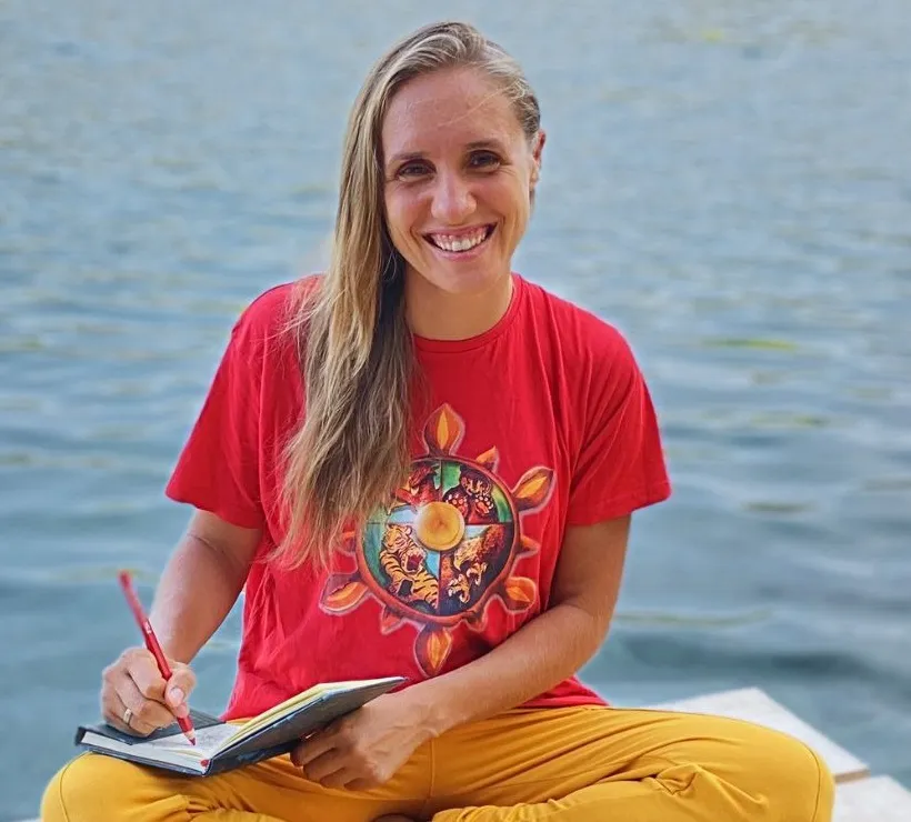 Santina sitzt lachend im Schneidersitz auf einem Holzsteg am Wasser, im roten SanTi-Shirt mit dem Rad des Kung-Fu, und schreibt in ein Notizbuch.