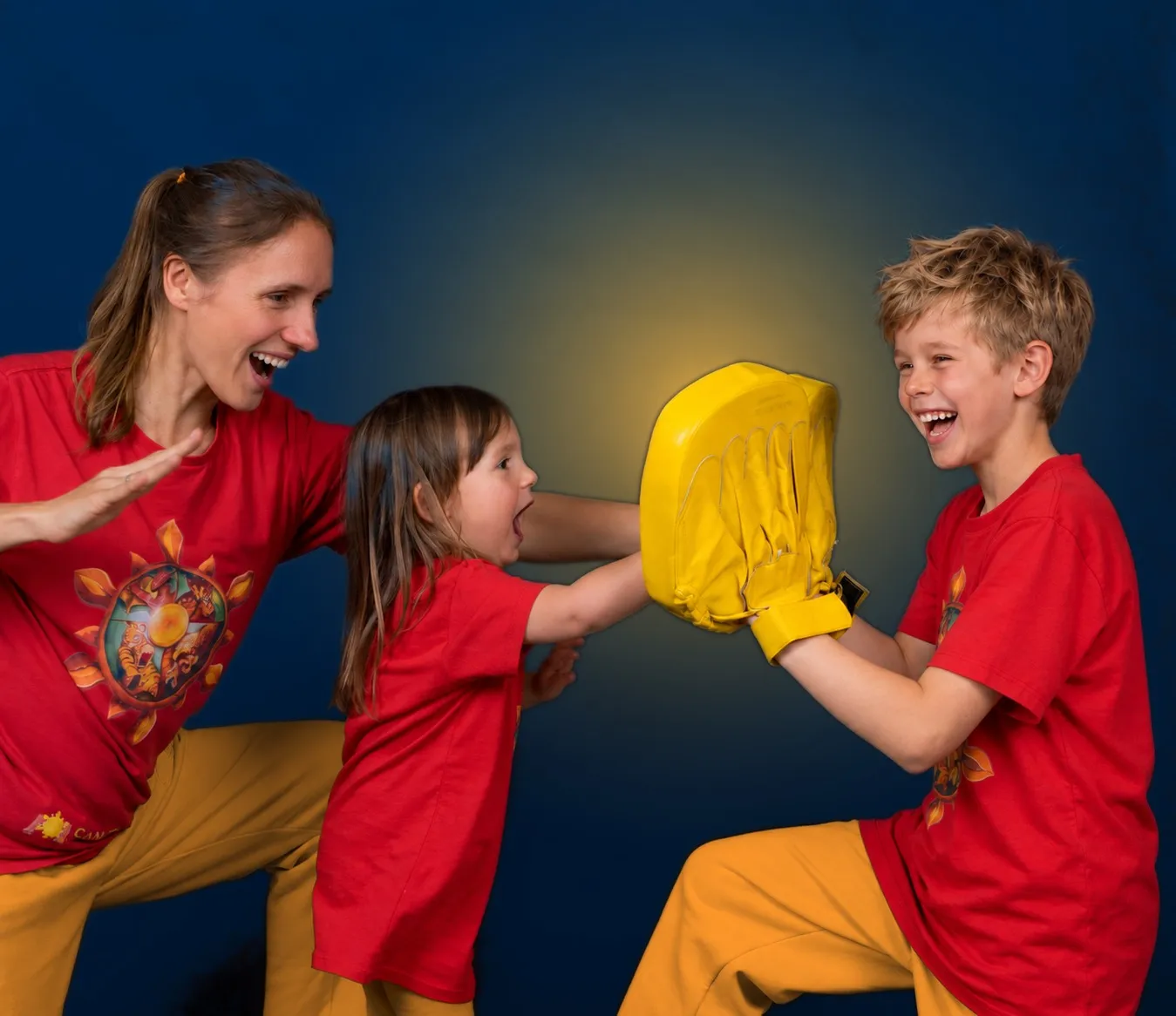 Santina übt lachend mit zwei Kindern in roten SanTi-Shirts: Das kleine Mädchen schlägt mit offener Hand auf eine gelbe Pratze, die der ältere Junge hält — alle drei lachen.